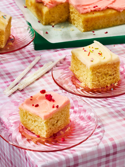 Square pieces of Glazed Cake Co Vanilla Bean and Lemon Raspberry cake mix cakes on a pink plaid tablecloth with Meri Meri gold glitter candles