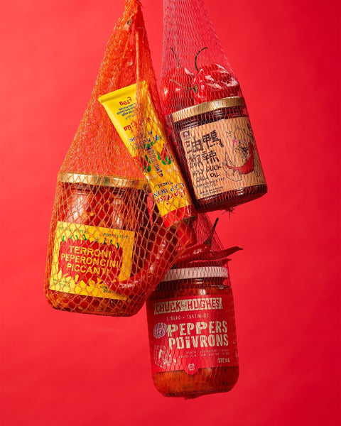 Red mesh bags holding Holy Duck chili oil, Terroni peperoncini piccanti, De Bon Cap Harissa and Chuck Hughes pepper spread against a red background.