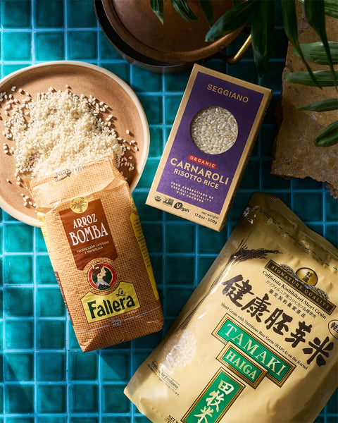 A bowl of La Fallera Bomba Rice with it's package and a box of Seggiano Organic Carnaroli rice and a bag of Tamaki Haiga rice on a blue tile surface