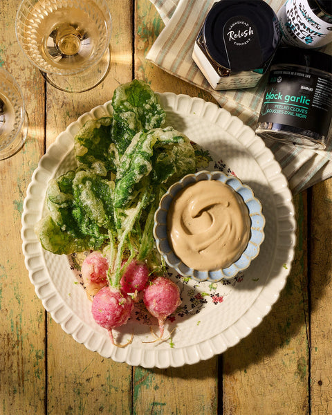 Radish tempura with black garlic aioli served on a white plate, with Hawkshead Black Garlic Relish and Silver & Green Black Garlic Oil in the background.