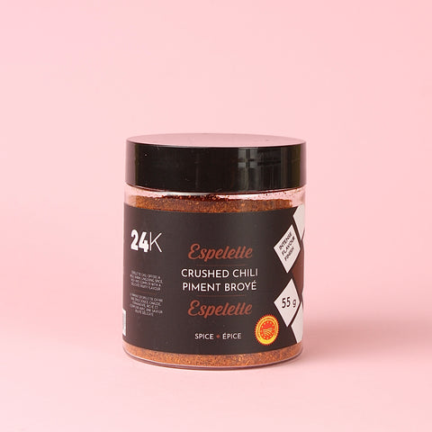 A jar of 24K Espelette Crushed Chili Piment Broyé spice, featuring a black lid and a black label with white and orange text, set against a solid light pink background.