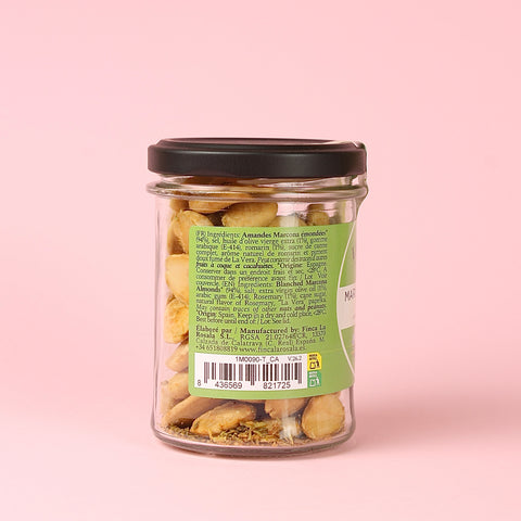 A clear glass jar filled with Finca La Rosala Marcona Almonds and Rosemary, sealed with a black lid, set against a solid light pink background. The back of the jar features a green label with text and a barcode.