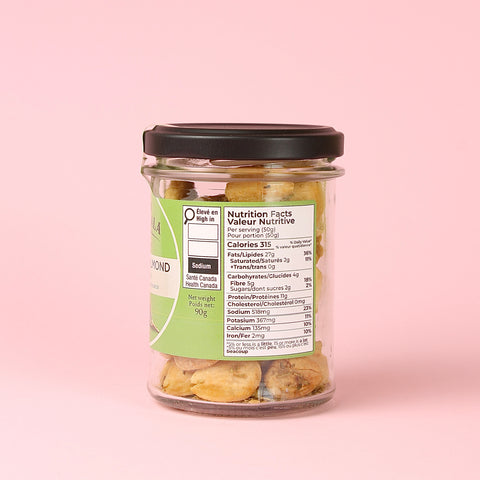 A clear glass jar of Finca La Rosala Marcona Almonds and Rosemary with a black lid and a green-and-white label showing nutrition facts, set against a solid light pink background