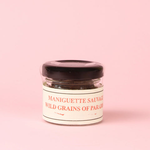 A small glass jar of Épices de Cru Maniguette Sauvage 'Wild Grains of Paradise' with a cream-colored label and a black lid, set against a solid light pink background.